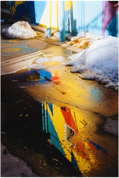 A film photograph of a winter sidewalk with melting snow and a dark puddle reflecting a brightly painted building in yellow, blue, and red. The wet pavement turns the reflection into abstract shapes, resembling mixed paint on a palette. Grain and soft focus emphasize texture and color rather than detail.