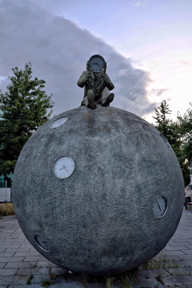 "Der Zeitzähler," a public sculpture by Gloria Friedmann in Magdeburg, features a large, textured grey sphere embedded with eighteen clock faces, of which five are visible here. Atop the sphere sits a bronze figure of a man in a suit, holding a clock in front of his face, symbolising the passage of time. The artwork is set in an outdoor environment, with trees and a cloudy sky in the background.