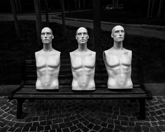 This black and white photo is of three pale, bald, male mannequins on a wooden park bench. (In Piazza San Francesco) Each mannequin is a torso, cut off below the waist. The mannequins' faces have neutral expressions, staring straight ahead. The bench is dark and worn, with visible text inscribed on the wood. The ground beneath the bench is paved with small, square cobblestones laid in a pattern. In the background, there are blurred trees and a path.