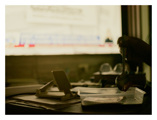 A cluttered desk featuring a hand holding a spoon over a plastic container. A wooden phone stand sits on some papers nearby, silhouetted against a bright, out-of-focus screen in the background