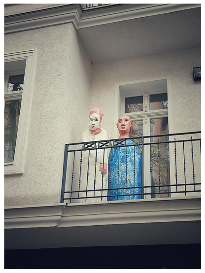Zwei lebensgroße Figuren auf einem Balkon, eine hält sich eine weiße Theatermaske vor das Gesicht.