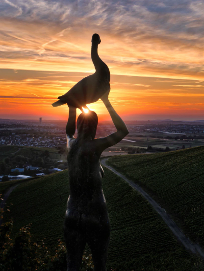 Die Silhouette einer Skulptur, die in den über Kopf erhobenen Händen eine Gans trägt.
Der Blick geht weit ins Tal gegen den Sonnenuntergang.