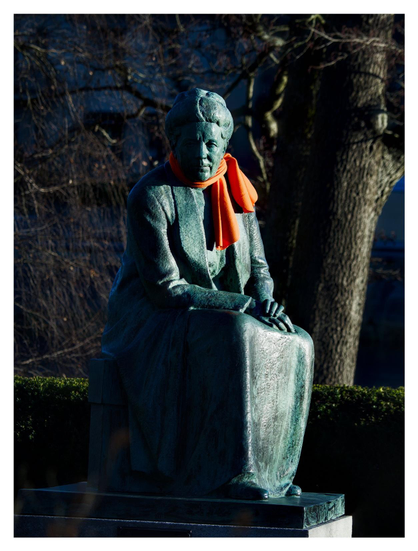 En stående bild på en staty av Selma Lagerlöf som sitter på en stol och tittar in i kameran. Hon har en orange halsduk. I bakgrunden ett träd och en grön häck.