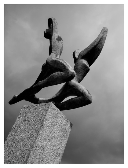 En stående svartvit bild på en staty på en piedestal i betong. Statyn är en man och en kvinna som sträcker sig mot himmelen som här är molnig.