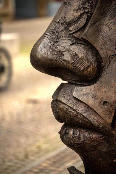 Eine Nahaufnahme in Seitenansicht zeigt die rechte Hälfte des Gesichts einer großen Bronzeskulptur. Die Skulptur hat eine dunkle, patinierte Oberfläche mit rauer, geformter Textur, die Falten, Risse und Narben im Material simuliert. Der Fokus liegt scharf auf der Nase und den Lippen. Die Nase ist groß und prominent, mit detaillierten Nasenflügeln. Die Lippen sind dick und leicht geöffnet, was dem Gesicht einen ausdrucksstarken Charakter verleiht. Im Hintergrund, der unscharf ist, ist ein gepflasterter Boden und links ein Teil eines Fahrradreifens zu erkennen, was auf einen öffentlichen Außenbereich hindeutet. Die Beleuchtung betont die reliefartigen Strukturen der Skulptur.