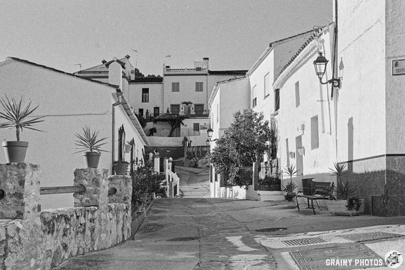 A black-and-white image of a quaint village street in Cartajima lined with whitewashed buildings, potted plants, and a bench. The path leads upward, showcasing a serene atmosphere and charming architecture typical of a Spanish village