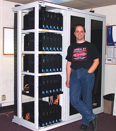 A person (man) in an "Area 51" T shirt stands next to a large server rack filled with many of networked Sony PlayStation 2 (PS2) consoles. 