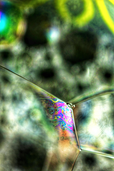 Die Aufnahme zeigt eine detaillierte Makroansicht von Seifenblasen, wobei der Fokus auf einem spezifischen, eckigen Schnittpunkt von drei Blasenmembranen im unteren Zentrum liegt. Dieses Segment weist intensive, wirbelnde Muster in leuchtenden Farben wie Magenta, Cyan, Gelb, Orange und Grün auf, die durch die Interferenz des Lichts mit der dünnen Seifenschicht entstehen. Die umgebenden Bereiche sind unscharf und zeigen eine Mischung aus dunkleren, körnigen Texturen und helleren, diffusen Flecken, die weitere Seifenblasen im Hintergrund andeuten. Der obere linke Bereich enthält einen hellen, regenbogenfarbenen Lichtfleck. Die Gesamtästhetik ist abstrakt und betont die komplexen, fließenden Farbmuster auf den transparenten Oberflächen.
