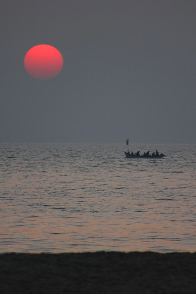 das bild zeigt einen sonnenuntergang über dem meer. der himmel ist in einem trüben grau, während die sonne, ein großer, roter ball, hoch oben am himmel steht. eine silhouette eines kleinen bootes mit einer gruppe von menschen an bord ist auf dem wasser zu sehen. das wasser spiegelt einen leichten rosafarbenen schimmer der sonne wider. im vordergrund ist ein dunkler strandstreifen zu erkennen. die szene wirkt ruhig und friedlich.