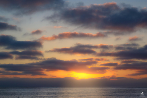 Color photo of a sunset sky filled with bands of clouds above the Pacific Ocean. 