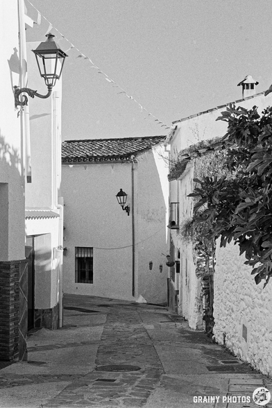 A narrow, cobblestone alleyway lined with whitewashed buildings and vintage lanterns, leading into the distance under a clear sky.