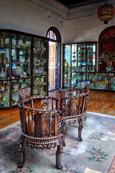 A pair of intricately carved tête-à-tête wooden chairs, adorned with mother-of-pearl inlays, takes centre stage in this image from the Pinang Peranakan Mansion in Malaysia. The chairs are elegantly placed on a floral-patterned rug, inviting intimate conversation. The room is richly decorated with glass display cabinets filled with ornate porcelain and ceramic artefacts, including vases, plates, and bowls, reflecting the opulent Peranakan heritage. The space is bathed in natural light, and a decorative hanging lantern enhances the cultural ambiance.