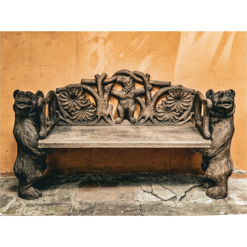 A rustic wooden bench with intricate carvings up against a textured orange wall. The bench is the focus, with two carved bear figures serving as the armrests. These bears stand upright, their mouths open in a roaring expression. The backrest of the bench has more carvings, including floral motifs and a central figure of a bear standing upright, holding onto wood carved branches.The wall behind the bench is a warm orange. The ground beneath the bench is paved with stone.