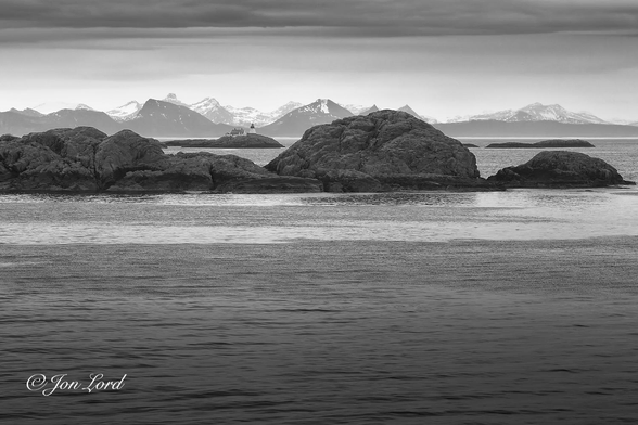 This is a black and white seascape photo of a remote arctic lighthouse in landscape format. Vestfjorden, Norway (2023).

In the foreground and rising upwards to the 2/3 up level is a body of rippled sea water. At this point is a small, undulating and rock islet stretching across the photo from left to right. Through a gap between the rocks is a distant view, about 500m, across further water of a small coastal lighthouse located on a small rocky island that's no more that 30 metres long. The white lighthouse is conical with a broad base and pointed top. To the left are 2 small white single story buildings, possibly the former lighthouse keepers cottage and a storehouse? In the background and a further, say 10km away, is a long range of jagged, snow covered mountains located on the coast of the mainland. 

The lighthouse is the Moholmen Lighthouse located in the middle of the Vestfjorden in Norway's arctic north.

https://en.wikipedia.org/wiki/Moholmen_Lighthouse