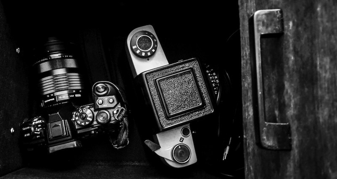 Man sieht eine geöffnete Schublade. Darin befindet sich eine digitale Olympus-Kamera wie auch eine analoge Mittelformat-Kamera vom Typ Kiev 60. Das Foto ist in Schwarzweiß.