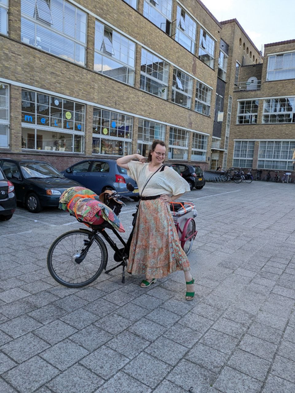 Elise is striking a pose in front of her bicycle and cart, which are both covered in items. She's wearing green heels under a colorful patterned orange skirt.