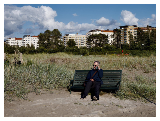 En liggande bild på en man som sitter på en grön träbänk och pratar i telefon. Framför är torkad lera, bakom högt grönt gräs och längst bort en rad av vita flerfamiljshus. Himmelen är blå med mestadels vita moln.