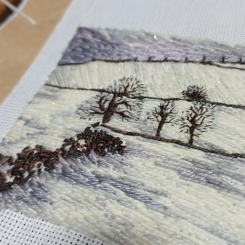 Part of a stitched artwork depicting a snowy landscape. A tumble down stone wall in the foreground with three bare trees and a bush behind. Snow covered fields and stone wall to the horizon. Viewed on an angle to show texture