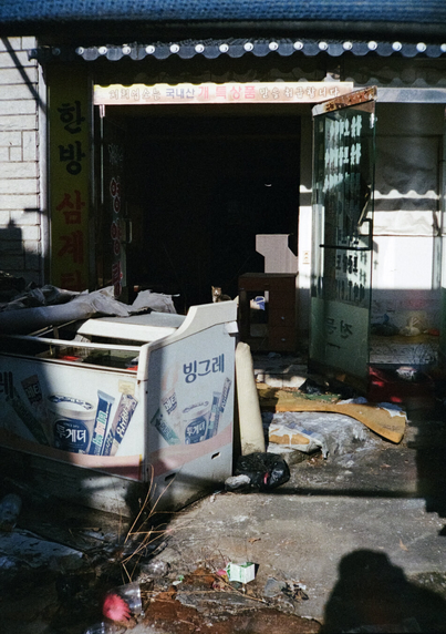 폐점한 지 오래되어 아이스크림 냉장고 등 각종 쓰레기로 가득한 식당. 식당 건물 안쪽에 고양이 한 마리가 카메라를 바라보고 있다.