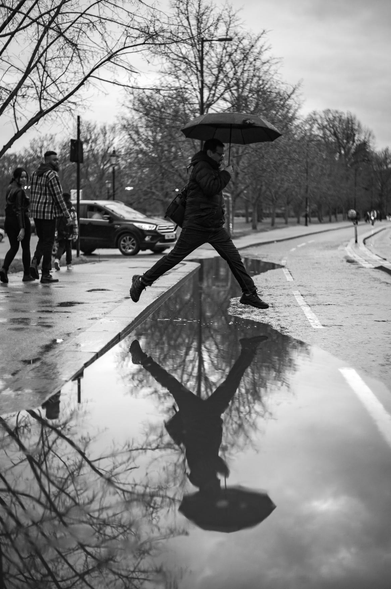 The image is a black and white, high-contrast photograph capturing a dynamic moment on a wet city street, on an overcast day. The main subject is a man in mid-air, leaping over a large puddle with an umbrella held above his head. His body is contorted, with his left leg extended forward and his right leg bent behind him. He is wearing dark clothing, including a jacket, pants, and athletic shoes, and he has a bag slung over his shoulder. The umbrella, also dark, is open and held by his right hand.