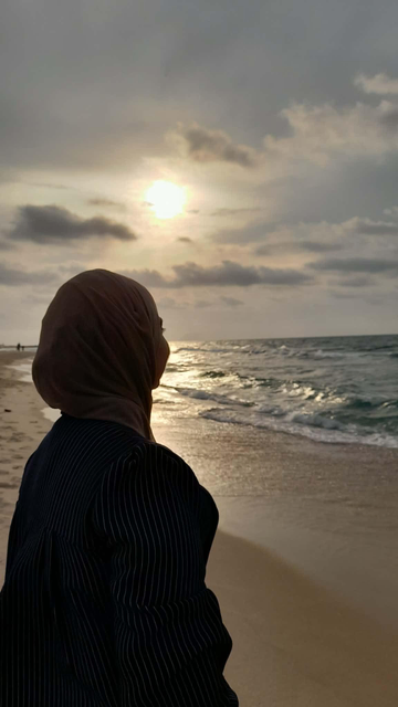 Naeema in a hijab looking at the ocean at sunset, sun reflecting on the water, wearing a striped blue shirt.