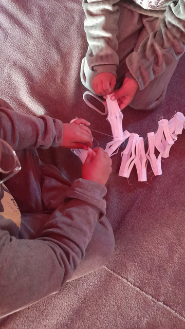 Two children, the twins Elias & Aylan, in grey pajamas making paper cut-out ornaments, sitting on a fluffy grey blanket. The children are focused on cutting and threading the paper.