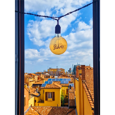 A photograph of a cityscape (Bologna) viewed through an open window. A yellow incandescent light bulb with "love" written in black cursive hangs by a blue cord against a bright blue sky with white clouds. Below there are yellow buildings with red-tiled roofs and solar panels.
