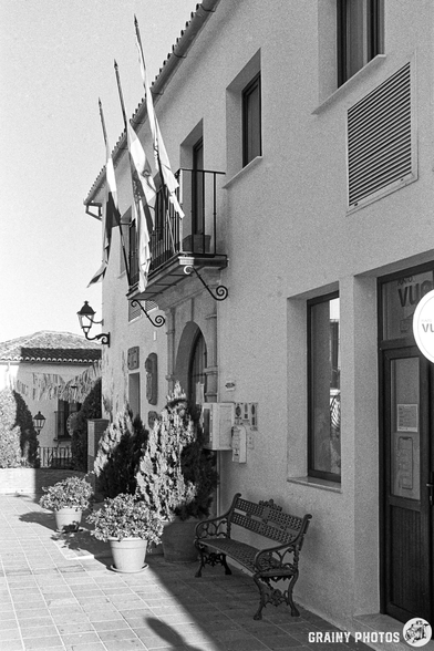 A quaint street scene featuring the Town Hall adorned with flags, a balcony, and a small bench. Potted plants flank the entrance, which has a shop sign visible on one side. The image is in black and white, capturing the charm of the architecture.