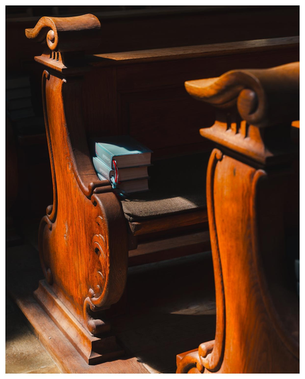 Bänke in einer Kirche mit Gesangbuch drauf. Viel Kontrast aus hellen von der Sonne beschienenen Partien und Schatten.