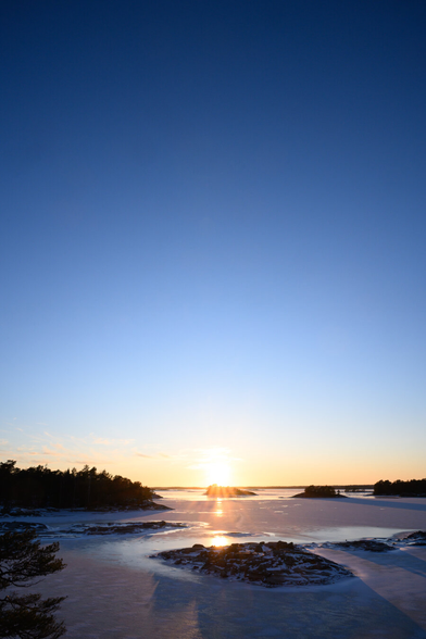 A vertically oriented landscape photography of the sun setting over a frozen over archipelago. The sky is taking up the vast majority of the vertical space in the photo, showing a gradient from yellow/orange at the horizon, to light blue, to deep darker blue at the top edge. The sky is entirely clear, apart from some very small, sparse clouds near the horizon.

The archipelago consists of a frozen over sea with just a little bit of snow on the ice, and a number of small rocky islands. The islands toward the horizon are predominantly covered by trees and the ones nearer the foreground are bare rock.

The sun is in the center of the frame, just above the horizon, casting a pale golden light over the landscape and creating deep shadows where it shines on the islands. There are golden reflections in the ice from the setting sun.