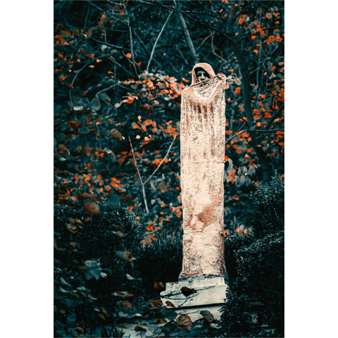 A photonof a tall, narrow statue of a figure draped in a robe, positioned in the centre foreground. The figure's head is covered by a cowl, obscuring her face, and she appears to be holding a piece of the robe in her hands, partially revealing the face, and draped across the front. The statue is made of a light-coloured stone and shows signs of age and weathering. The statue stands on a tiered base, which is also weathered. Behind the statue, there is a dense collection of trees and bushes with a mix of green and orange leaves.