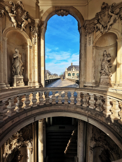 This image captures a breathtaking architectural view of the Zwinger Palace in Dresden, Germany. The photograph is taken from an ornate archway, framed by intricate Baroque sculptures and decorative balustrades. Two statues, one on each side of the arch, depict classical figures, adding to the grandeur of the scene.

Beyond the archway, a long, symmetrical pathway stretches into the distance, flanked by low walls and additional statues. The pathway leads the eye towards a stately building in the background, set against a vivid blue sky with scattered clouds. The interplay of light and shadow enhances the intricate details of the stonework, showcasing the elegance and opulence of Baroque architecture.