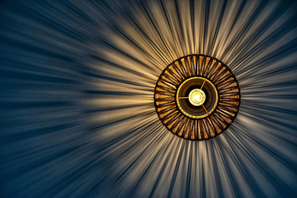 Das Bild zeigt eine kreisförmige Deckenleuchte in einer Nahaufnahme von direkt unten, die ein spektakuläres Licht- und Schattenspiel an die Wand wirft.
Im Zentrum der Komposition befindet sich die leuchtende Glühbirne, die ein warmes, gelbliches Licht ausstrahlt. Sie ist von einem runden Lampenschirm umgeben, der aus vielen dünnen, dunkelbraunen oder schwarzen Stäben besteht, die strahlenförmig von der Mitte nach außen verlaufen. Diese Stäbe sind durch zwei konzentrische Ringe miteinander verbunden, was dem Schirm eine strukturierte, fächerartige Optik verleiht.
Das markanteste Merkmal ist der dramatische visuelle Effekt: Das Licht der zentralen Quelle dringt durch die Zwischenräume der Stäbe und erzeugt hunderte von feinen, hellen Lichtstrahlen, die sich symmetrisch und weiträumig über die gesamte dunkelblaue Wandfläche ausbreiten. Zwischen diesen Lichtstreifen entstehen ebenso feine, dunkle Schattenlinien, wodurch ein dynamisches Muster entsteht, das an eine strahlende Sonne oder eine Explosionszeichnung erinnert. Die tiefblaue Farbe der Wand bildet einen starken Kontrast zum warmen, goldenen Schein des Lichts und verstärkt die Tiefenwirkung des Musters.