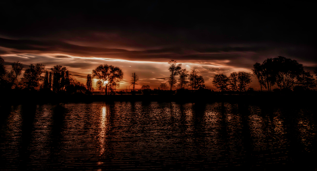 Man sieht einen fast kupferfarbenen Sonnenuntergang an einem kleinen See. Die Sonne scheint durch einen Baum und spiegelt sich auf der nicht ganz ruhigen Wasseroberfläche wieder. Weitere Bäume und Sträucher sind am gegenüber liegenden Ufer zu erkennen. Oberhalb der untergehenden Sonne sieht man dunkle Wolken die die Nacht ankündigen.