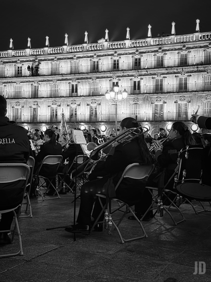 La imagen muestra una banda de música tocando al aire libre, sentada en sillas plegables, con instrumentos de viento y percusión. Al fondo se ve un edificio histórico iluminado, con varias plantas, balcones y una barandilla en la parte superior. La escena es nocturna y está capturada en blanco y negro, transmitiendo un ambiente solemne y elegante.