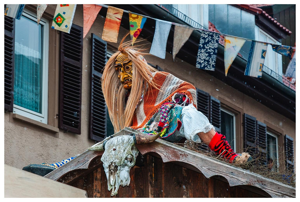 Hexe mit Holzmaske und gestricktem Tuch sitzt auf einem Wagen, der wie ein Hexenhaus aussieht.