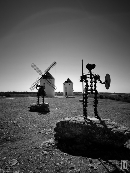 La imagen en blanco y negro muestra un paisaje rural con varios molinos de viento tradicionales. En primer plano, se ven dos esculturas metálicas que representan a Don Quijote y Sancho Panza, situadas sobre rocas. Al fondo, destacan los molinos bajo un cielo despejado, creando un contraste marcado entre las siluetas oscuras de las figuras y la luminosidad del horizonte.
