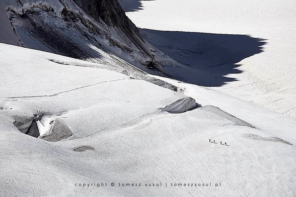 This aerial view shows a pristine glacial landscape, featuring a vast expanse of white snow and ice with subtle textural patterns caused by wind and snow movement. Smooth, undulating snowfields dominate the foreground, showing gentle ridges and natural flow lines. In the middle distance, dark rocky outcrops and crevasses break through the white surface, creating a stark contrast, including two prominent, oval-shaped holes revealing dark rock beneath. The upper part of the image shows steep, dramatic mountain cliffs with exposed dark grey and brown rock faces, which create sharp diagonal lines across the frame. Four tiny dark silhouettes representing human figures are visible in the lower right quadrant of the snowfield, providing a sense of scale to the vast landscape. Strong shadows are cast along the cliff edges and in the crevasses by the lighting, emphasising the dramatic topography and three-dimensional relief of this high-altitude glacial environment.