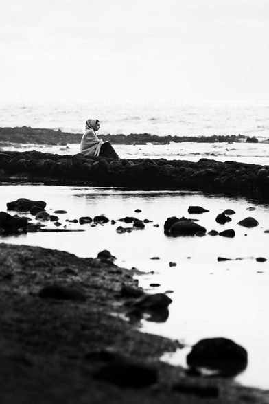 A monochrome photo of a person covered by a blanket sitting at a beach. A pool of water between the foreground and the person.