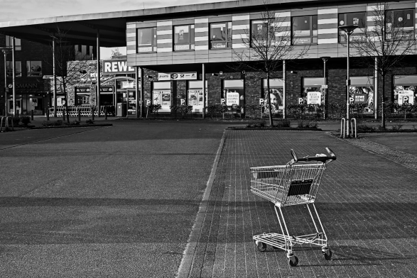 Ein einsamer Einkaufswagen steht vergessen auf dem Parkplatz eines Supermarktes. Im Hintergrund das Gebäude des Supermarktes, auf dessen linker Seite vor dem Eingang viele weitere, gesammelte Einkaufswagen stehen. 
