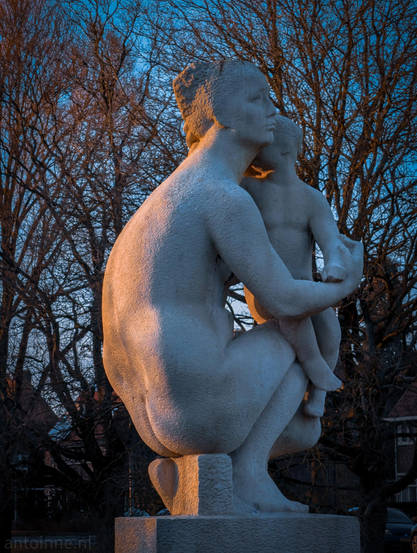 A stone sculpture of a mother and child, set against a backdrop of bare, winter trees and a deep blue twilight sky. The warm, golden light of a setting sun hits the side of the figures, creating a high-contrast "golden hour" effect that highlights the texture of the stone.

The sculpture (by Joop Hekman) depicts a woman in a deep crouch, cradling a young child against her chest. Her posture is protective and grounded. The child leans into her, emphasizing a sense of security and intimacy.
