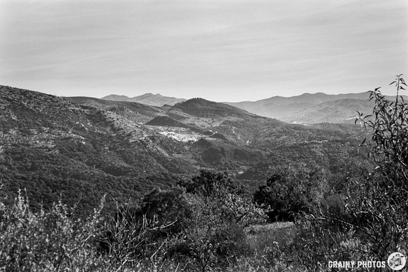 A panoramic view of rolling hills and mountains, captured in black and white. The landscape features dense vegetation and varying elevations, with a serene atmosphere and subtle layers of mist in the distance.