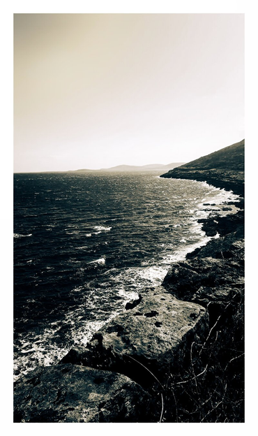 The photograph is of Blackhead in co.Clare. It is a black and white scene with waves from the ocean that stretch towards a distant horizon under a pale sky. The water, is in varying shades of grey and helps show their texture. The coastline is defined by dark, rugged rocks and cliffs, their surfaces uneven and textured, contrasting against the sea. In the distance, the hazy silhouettes of hills are barely visible on the horizon.