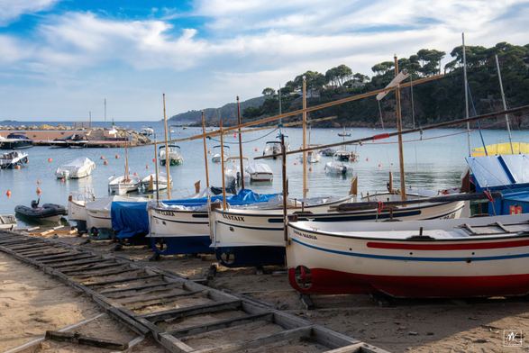 Color photo of small boats hauled up on a beach with a wooden trackway for them in the foreground and extending down towards the water. In the background is a small harbor with boats on the water and beyond that is a tree covered, hilly shoreline extending into the distance. 
