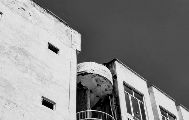 A low-angle, black-and-white view of the upper façade of a weathered building against what-would-have-been a bleak blue sky. A tall textured wall on the left contains two small, dark, square windows, while the section to the right features larger rectangular windows. Nestled between these sections is a rounded semi-circular balcony structure that appears to have peeling paint and plaster on its underside, sitting above a curved metal railing. The building's surface is rough and white-colored, contrasting sharply with the deep color of the sky. c0e238a3-4964-46d4-91c9-2157efb1ee58