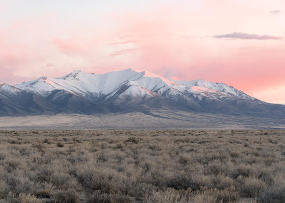 A color landscape photo of a tall snow-capped distant mountain in a vast flat plain of sagebrush. There are a few clouds in the sky, especially on the left, showing red-orange sunset colors. Some of the color is reflected in the white snow on the mountain top.