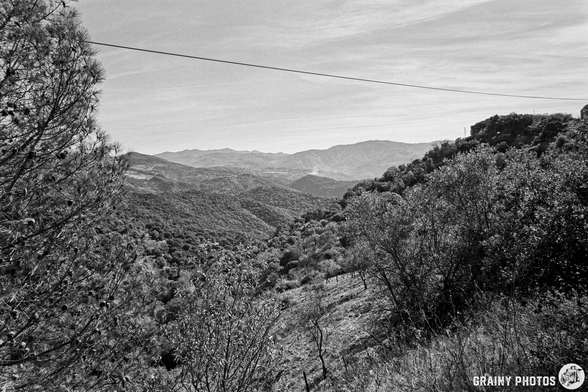 A black and white landscape showing rolling hills and mountains in the distance, dotted with trees and shrubs, under a lightly clouded sky. A power line stretches across the top of the image.