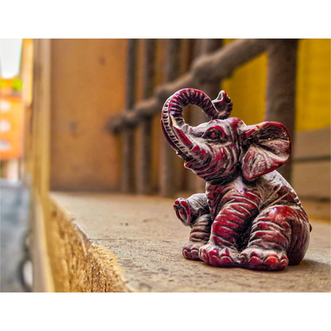 The photograph is of a reddish-brown elephant figurine placed on a weathered, light-brown ledge. The elephant, detailed with carved lines and a slightly glossy finish, is sitting upright with its trunk raised. The background is intentionally blurred, focussing on the elephant. Vertical bars and a muted yellow wall are visible in the background.The overall lighting is soft and natural, casting gentle shadows around the figurine.