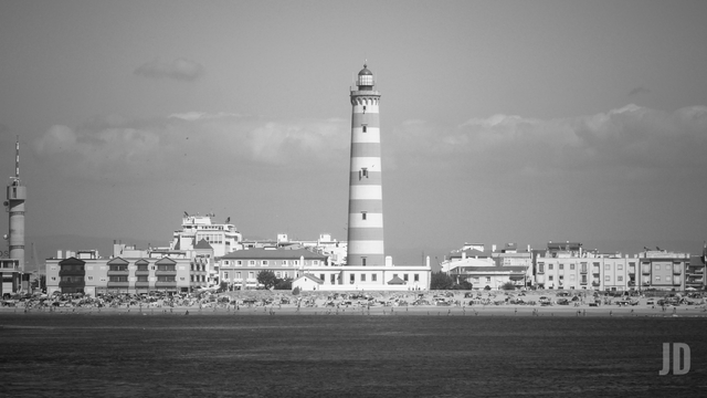 La imagen muestra un faro alto con franjas horizontales, situado junto a una playa poblada de sombrillas y bañistas. Detrás del faro se extiende un conjunto de edificios de baja y media altura, bajo un cielo parcialmente nublado. La foto está en blanco y negro, lo que resalta el contraste entre el mar en primer plano y el horizonte urbano.