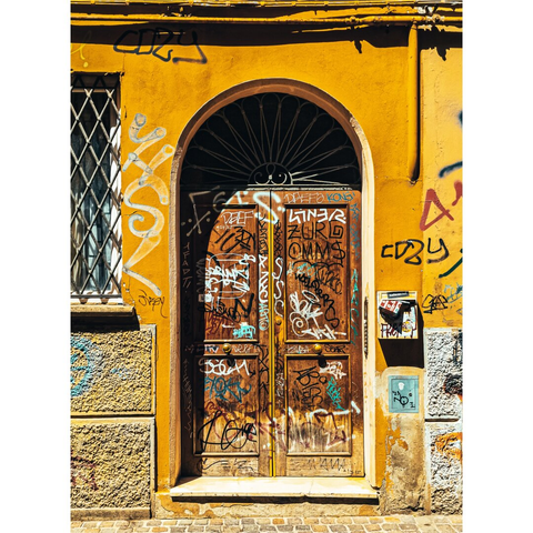A photograph of a weathered, wooden double door set in a vibrant yellow, graffiti-covered wall. The door is adorned (a-door-ned) with numerous coloured tags, including names and symbols. To the left, a window with a diamond-patterned metal grille is partially visible. The wall has textured, grey/beige stone blocks at the bottom. Bright sunlight casts sharp shadows.